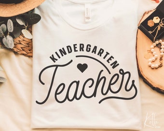 May include: White t-shirt with black text that reads "Kindergarten Teacher" in a script font with a small black heart above the word "Teacher".
