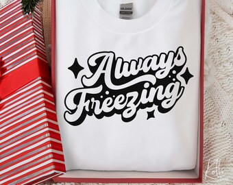 以下が含まれることがあります： 白いスウェットシャツに、レトロなフォントで「Always Freezing」という黒い文字が書かれています。文字の周りは小さな星で囲まれています。