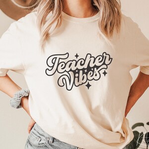 Puede incluir: Una camiseta blanca con el texto "Teacher Vibes" en una fuente retro con estrellas negras.