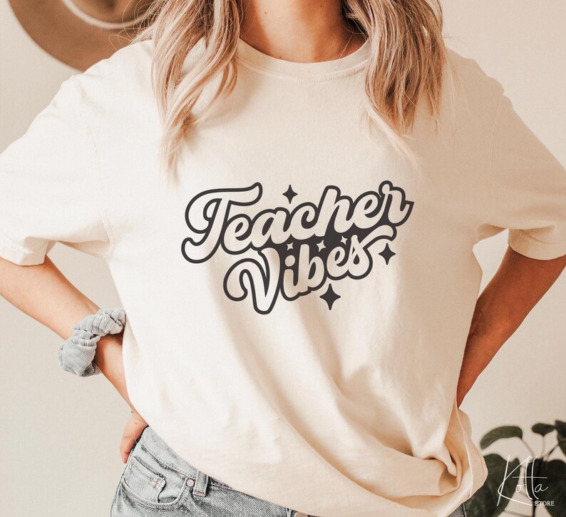 Puede incluir: Una camiseta blanca con el texto "Teacher Vibes" en una fuente retro con estrellas negras.
