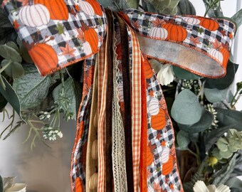 May include: A decorative bow featuring a black and white buffalo check pattern with orange pumpkin and autumn leaf prints. The bow includes long, cascading ribbons in various textures, including burlap, orange gingham, and dark brown velvet.