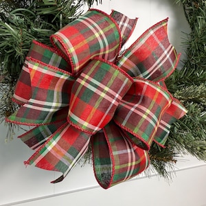May include: A festive Christmas wreath featuring a large plaid bow in red, green, and brown. The bow is the focal point, with a classic tartan pattern and red trim. The wreath is made of artificial evergreen branches.