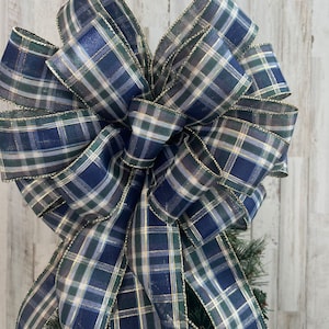 May include: A large plaid Christmas tree topper bow in navy blue, green, and white with gold accents. The bow is made of wired ribbon and has a loop at the top for hanging.