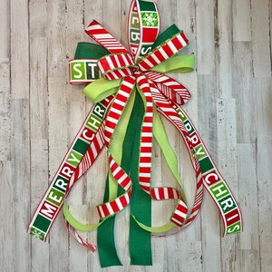 May include: A festive Christmas bow with red and white striped ribbon, green and lime green ribbons, and the words "MERRY CHRISTMAS" in green and white. The bow features a snowflake design and is perfect for holiday decorating.