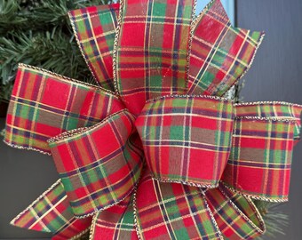 May include: Close-up of a decorative bow with a classic plaid pattern in red, green, and gold. The bow is made of fabric and has a gold trim. The bow is likely used for holiday decorations.