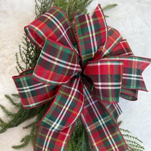 May include: A large, decorative bow made of red, green, and white plaid ribbon. The bow is accented with a red trim and is attached to a bed of green pine needles. The bow is likely for holiday or seasonal decor.