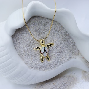 May include: A gold-toned turtle pendant necklace featuring an iridescent white opal inlay on the shell. The turtle charm is suspended from a thin gold-toned snake chain, showcasing a polished metallic finish and a detailed sea creature design.