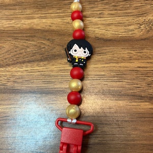May include: A red and gold silicone beaded clip with a Harry Potter character charm. The clip is attached to a white string.