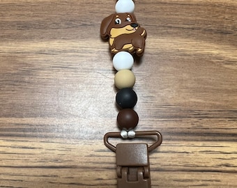 May include: A brown silicone baby teether with a brown and white dog charm. The teether has a brown clip and is strung with brown, black, and white beads.