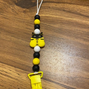 May include: A yellow and black silicone baby teether with a bee design and a yellow clip. The teether has a white string attached to it.