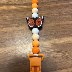 May include: A silicone teether with a butterfly charm and a clip. The teether is orange, white, and cream colored.