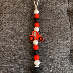 May include: A baby pacifier clip with a red tractor charm. The clip features a string of red, black, and white beads. The tractor charm is red with black wheels. The clip has a white cord loop and a black plastic clip.