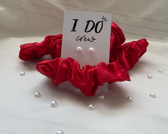 May include: A white card with the text "I Do Crew" on it. Two small pearl earrings are displayed on the card. The card is sitting on top of a red satin scrunchie.