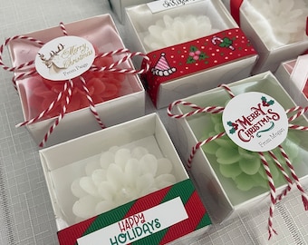 May include: Assortment of small, clear gift boxes with white, green, and red candles. Each box is decorated with a red and green ribbon, a white circle sticker with a holiday message, and a personalized message. The boxes are arranged on a white and gray checkered surface.