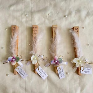 Puede incluir: Cuatro abanicos de mano de madera con plumas blancas, flores y etiquetas que dicen "Stay Cool!". Cada abanico tiene una flor con una perla en el centro y una pequeña etiqueta. Los abanicos están dispuestos sobre una superficie crema.