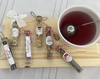 May include: Six small glass tubes filled with different types of tea blends. Each tube has a festive Christmas-themed label with a red and green color scheme. The labels include the text "Merry Christmas" and "Happy Holidays". The tubes are arranged on a wooden surface next to a white mug filled with red tea.