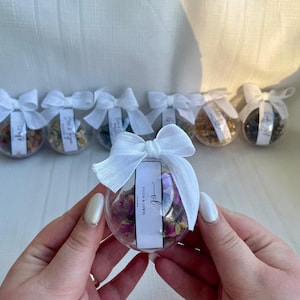 Puede incluir: Una colección de pequeños adornos de vidrio transparente llenos de flores secas y atados con una cinta blanca. Cada adorno tiene una pequeña etiqueta blanca con el texto "Viola & Lewis" y un "tel" escrito a mano.