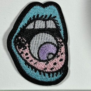 Peut inclure: Un patch brodé représentant une bouche grande ouverte avec un œil à l'intérieur. L'œil est entouré de broderies roses et noires, et la bouche est bordée de bleu.