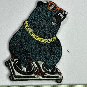 Könnte beinhalten: Gestickter Aufnäher eines schwarzen Bären mit Sonnenbrille und einer goldenen Kette, der hinter einem DJ-Mixer steht.
