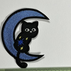 Peut inclure: Chat noir assis sur une lune croissante bleue avec des étoiles dorées et bleues. La lune est bordée d'un fil noir.