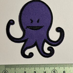 Peut inclure: Patch brodé d'une pieuvre violette avec un visage souriant et deux bras levés. Le patch mesure environ 5 pouces de large.
