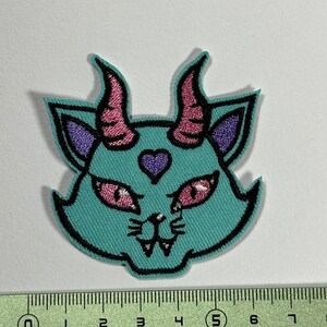 Puede incluir: Un parche bordado de un gato con cuernos rosas, orejas moradas y ojos rojos. El gato es azul turquesa y tiene un pequeño corazón morado en la frente.