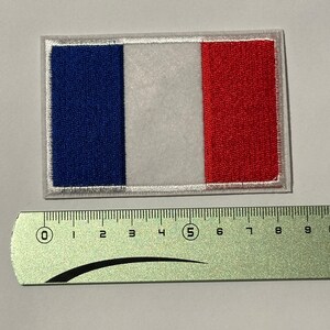 Peut inclure: Un écusson rectangulaire représentant le drapeau français. Il présente des bandes verticales bleues, blanches et rouges, avec une bordure blanche. Une règle est placée en dessous, indiquant les mesures en centimètres.