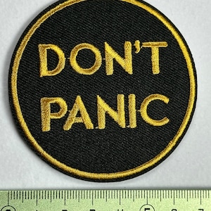 Könnte beinhalten: Ein runder schwarzer Patch mit goldener Stickerei. Der Text "DON'T PANIC" ist in Gold gestickt.