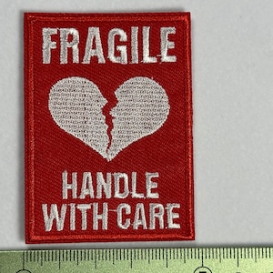 Peut inclure: Patch en tissu rouge avec broderie blanche qui indique "FRAGILE" avec un symbole de cœur brisé et "MANIPULER AVEC SOIN".