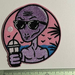 Peut inclure: Patch brodé d'un extraterrestre portant des lunettes de soleil et tenant une boisson, sur un fond rose avec des palmiers et des oiseaux.