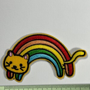 Peut inclure: Patch brodée thermocollante représentant un chat jaune avec un arc-en-ciel derrière lui. Le chat a un design simple et cartoon avec des yeux noirs et un nez rose.