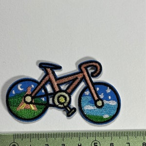 Op de afbeelding: Geborduurde strijkpatch van een bruine fiets met blauwe wielen. De wielen hebben een nacht- en daghemelontwerp met sterren, wolken en een halve maan.