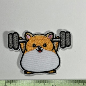 Peut inclure: Un patch brodé thermocollant représentant un hamster de dessin animé soulevant une barre d'haltères. Le hamster est brun et blanc avec une expression joyeuse. La barre d'haltères est grise.