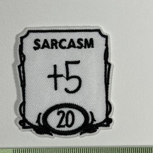Peut inclure: Écusson brodé blanc avec bordure et texte noirs. Le mot "SARCASM" est en haut, avec "+5" en dessous. Un ovale en bas porte le chiffre "20". L'écusson est sur une surface blanche à côté d'une règle.