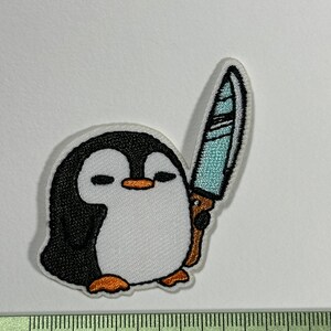 Peut inclure: Un pingouin blanc avec des ailes noires et un bec orange tenant un couteau bleu et blanc. Le pingouin est brodé sur un tissu blanc.