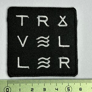 May include: Black square patch with white embroidered letters spelling "TRAVELER" with symbols in between each letter.