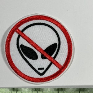 Peut inclure: Un écusson brodé blanc avec un cercle rouge et une ligne rouge diagonale qui le traverse. À l'intérieur du cercle se trouve un visage d'extraterrestre noir et blanc.