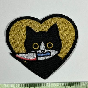 Peut inclure: Un chat noir avec des marques blanches tenant un couteau dans sa bouche, brodé sur un patch en forme de cœur doré.