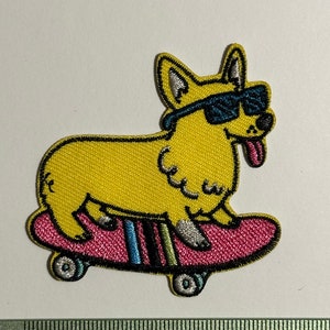 Puede incluir: Parche bordado amarillo de un perro con gafas de sol y haciendo skateboard. El perro está en una patineta rosa con rayas azules y blancas. Las ruedas son grises.