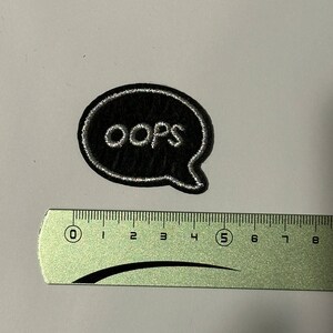 Peut inclure: Un écusson thermocollant brodé noir et blanc avec le mot "oops" dans une bulle de dialogue blanche.
