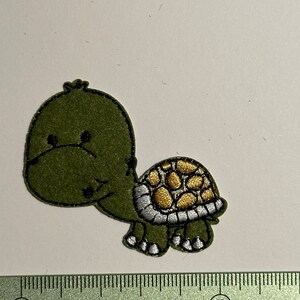 Könnte beinhalten: Ein grüner, schildkrötenförmiger Aufnäher mit einem lächelnden Gesicht und einem gold-silbernen Panzerdesign. Der Aufnäher ist etwa 5 cm lang und dient wahrscheinlich dekorativen Zwecken.