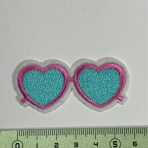 Könnte beinhalten: Ein türkisblauer herzförmiger Sonnenbrillen-Patch mit rosa Nähten und weißem Umriss. Der Patch ist etwa 5 cm breit.
