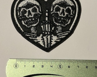 May include: Black and white embroidered patch of two skeletons in a heart shape. The skeletons are smiling and have their arms around each other. The patch is about 5 cm wide.