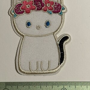 Peut inclure: Patch thermocollant brodé blanc représentant un chat portant une couronne de fleurs roses et violettes. Le chat a des yeux noirs et une queue noire.