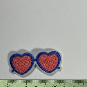 May include: A pink heart-shaped sunglasses patch with blue stitching. The patch is about 2 inches wide.