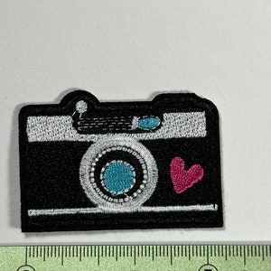 May include: Black and white embroidered camera patch with a pink heart. The camera has a blue lens and white details.