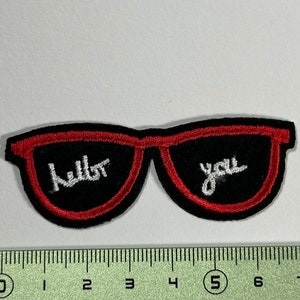 Peut inclure: Lunettes de soleil noires avec une bordure rouge et les mots "Hello You" brodés en blanc.
