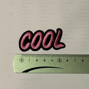 Peut inclure: Patch thermocollant brodé rose avec le mot "COOL" en lettres noires soulignées. Le patch mesure environ 5 cm de large.