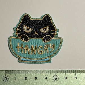 May include: A black cat with angry eyes peeking over the rim of a light blue bowl with the word "HANGRY" embroidered in gold thread.