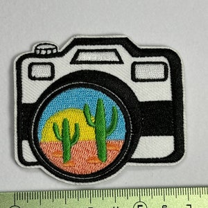 Peut inclure: Patch brodé blanc représentant un appareil photo avec un objectif noir. L'objectif montre une scène de désert avec un soleil jaune, un ciel bleu et deux cactus verts.